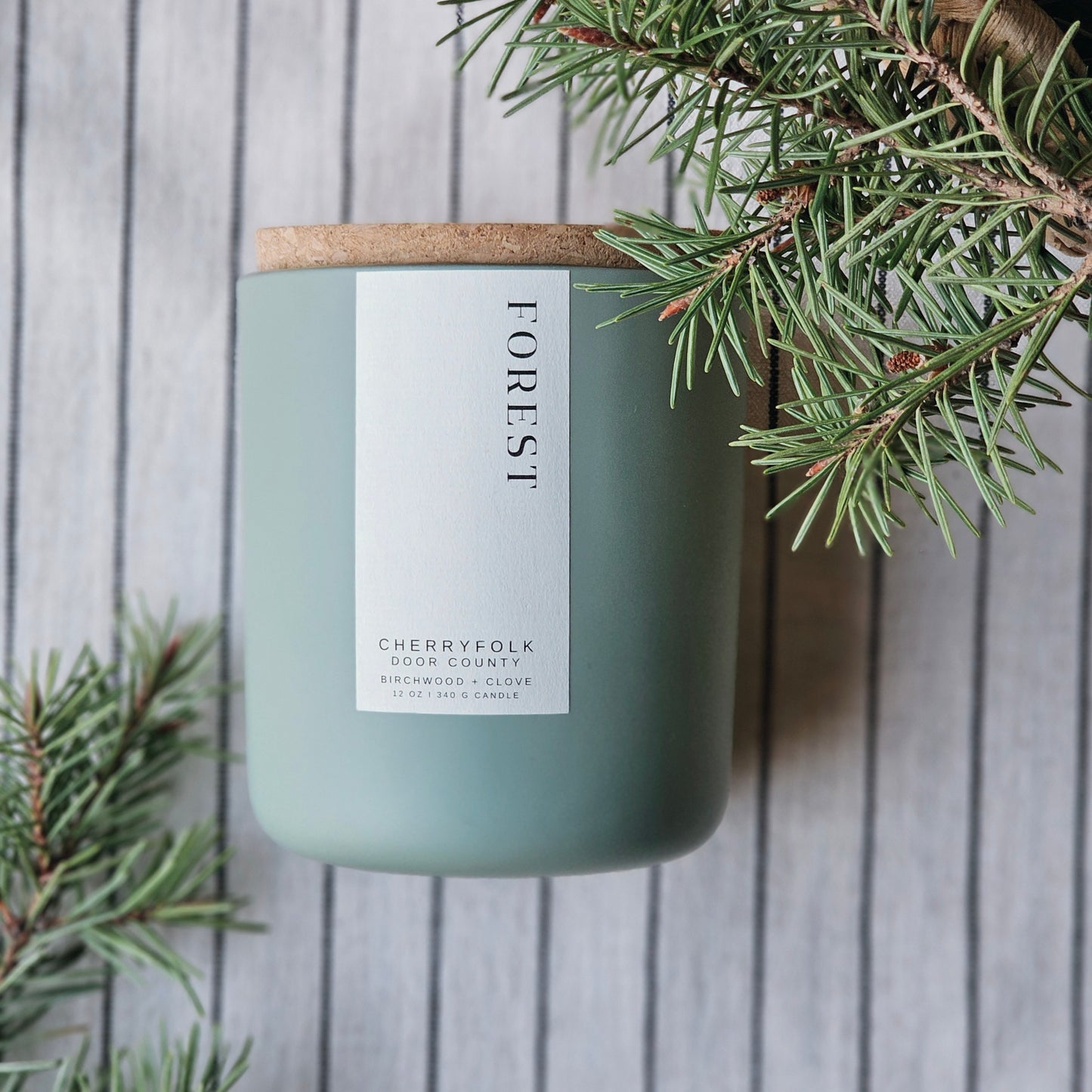 A matte sage green glass candle vessel with a cork lid labeled 'FOREST' containing a crackling wooden wick candle. Through the forest, a wanderer travels. A breath of needles, a touch of bark.